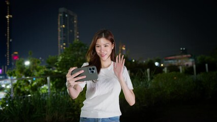 Wall Mural - Smiling woman taking selfie at night in city, waving joyfully amidst urban greenery and stunning buildings, capturing happiness in vibrant atmosphere. Asian Digital technology lifestyle