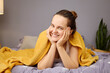 © sementsova321 - A woman lies on her stomach on a bed, her hands resting on her cheeks. She is wrapped in a soft yellow blanket and smiling cheerfully in a brightly lit, comfortable room