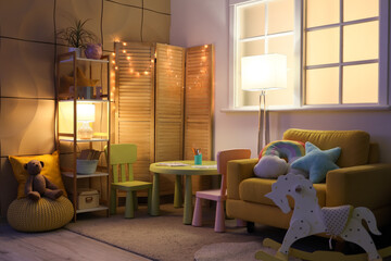 Wall Mural - Interior of stylish children's room with cozy armchair, toys, glowing lights and folding screen at night