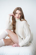 © alipko - young woman in a cozy beige oversized sweater and knee-high socks poses against a white wall. Her long, wavy hair frames her smiling face, exuding warmth and comfort in a minimalist setting
