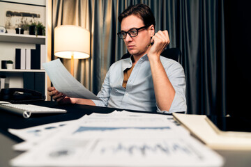 Wall Mural - Smart businessman holding paperwork report paragraph dynamic database, looking at pc screen with analyzing global target evaluation value profit company at modern home office at night time. Pecuniary.