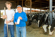 © JackF - Positive aged man dairy farm owner standing with teen grandson near stalls with cows on sunny summer day, holding bottle of fresh milk ..