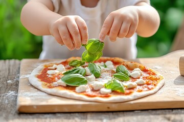 Wall Mural - Small child adding fresh basil leaves to a homemade pizza with tomato sauce and mozzarella cheese on a wooden table