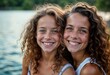 © miss irine - Two young girls sit by river smiling, embracing. Look happy, carefree. Curly brown hair, light skin. Portrait style photo. Outdoor scenery. Perfect for lifestyle magazines, family photos, social