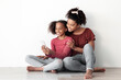 © Prostock-studio - Beautiful black mother and daughter using mobile phone together, sitting on floor, hugging and taking selfie for social media, enjoying time together at home, white empty wall backgound, copy space