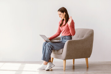 Wall Mural - Full length of millennial lady using laptop for online communication, sitting in armchair against white wall, blank space. Young Caucasian woman having video chat with friend, waving at camera