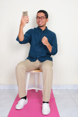 Wall Mural - A man sitting on the chair showing excited expression when looking at his phone