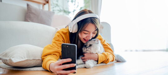 Wall Mural - Spending your free time at home with your cat. young asian woman in headphones, with mobile phone, lies on floor in living room, with fluffy Maine Coon cat, listens to music