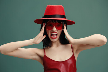 woman in red hat and sunglasses with hands on head and mouth open in surprise, against a green backg