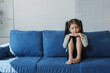 © Phimwilai - Young girl with pigtails sits on a blue sofa, looking contemplative and thoughtful. The bright natural light from the window highlights her peaceful expression in a minimalist setting