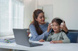 © Phimwilai - Mother assists her young daughter in adjusting headphones while sitting at a desk with a laptop. The setting suggests a cozy home environment, emphasizing family bonding and technology use
