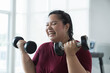 © amorn - Happy smiling Asian young plus size woman lifting dumbbell weight in fitness club. Young overweight woman holding dumbbell workout at gym. Asian woman exercise training with weight, healthy lifestyle