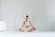 © yavdat - A young woman sits in a lotus position on a white floor, maintaining a calm and focused expression. This scene reflects the growing trend of mindfulness and self-care in modern urban lifestyles.