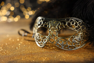 Beautiful carnival mask and black feathers on golden background against blurred lights, closeup. Bokeh effect