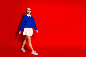 Wall Mural - Stylish young woman in blue sweater and white skirt walking confidently against vibrant red background