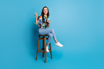 Wall Mural - Full length photo of smart woman wear print shirt sitting on bar stool hold smartphone show v-sign isolated on blue color background