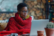 © Sascha - African woman wearing glasses working at home. Concept of comfort and relaxation, as the woman is wrapped in a warm blanket while working on her laptop