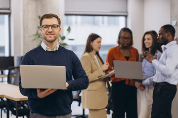 Wall Mural - Portrait of smiling businessman holding laptop with diverse business team collaborating on project in modern office