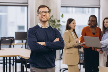 Wall Mural - Professional businessman confidently standing with crossed arms, colleagues collaborating in sleek corporate workspace