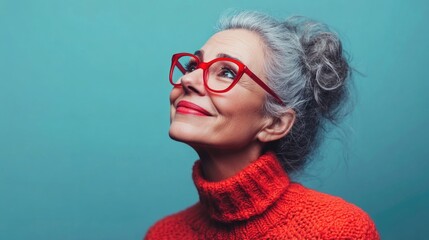 Wall Mural - Smiling mature woman with gray hair and red eyeglasses wearing a cozy orange sweater, posing against a teal background for advertising and lifestyle concept.