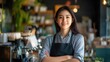 © The 2R Artificiality - Confident Asian woman entrepreneur smiling in coffee shop setting, wearing apron in cozy restaurant interior with plants and casual decor, showcasing successful small business ownership.