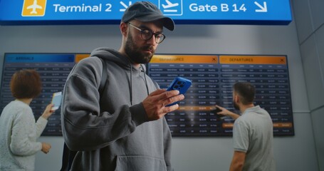 Busy Airport Terminal: Man with Backpack Checks Flight Information Scanning Ticket Using Phone. Traveler Going on Holiday Trip. Digital Arrival and Departure Board for Passengers In the Background.