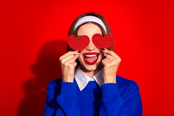 Wall Mural - Happy woman holding glitter hearts over her eyes against a red backdrop, dressed stylishly in a blue outfit with a white headband