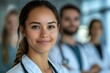 © Yuliia - Young female medical professional in white coat smiling at camera with healthcare team members standing in background, representing medical staff and teamwork.
