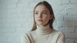 © The 2R Artificiality - Slender teenage girl with light brown hair wearing a beige turtleneck in natural light against a textured white brick wall showcasing natural beauty and skin imperfections