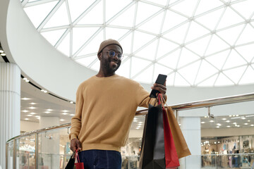 Wall Mural - Happy young consumer and blogger with paperbags using smartphone while standing against railings in the mall