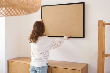 Wall Mural - Young woman hanging picture on white wall in stylish room