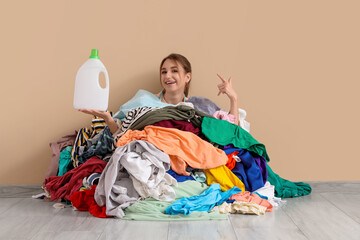 Wall Mural - Young woman with bottle of detergent buried under pile of dirty laundry on beige background