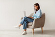 © Prostock-studio - Smiling young woman in casual wear working online, sitting in armchair and using laptop against white studio wall, copy space. Cheerful Caucasian lady surfing internet on portable pc