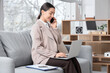 © Pixel-Shot - Young businesswoman with laptop talking by mobile phone on sofa in office
