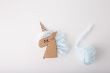 Carton unicorn head in center with light blue yarn ball on right side. White background, soft yarn texture. Step in creative process, top view. DIY unicorn craft using simple materials for projects.