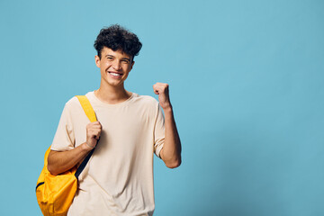 Wall Mural - Happy young man celebrating success with a cheerful smile, wearing a casual light shirt and a bright yellow backpack against a solid blue background