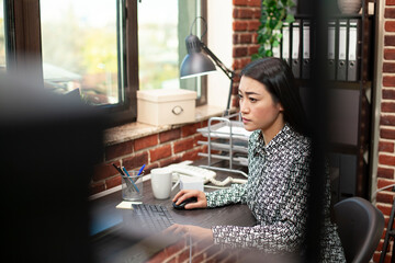 Wall Mural - Female researcher near window types diligently, updating marketing analytics in brick wall office. Asian woman focuses on project updates at her desk, working on her computer in a modern workspace.