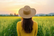 © SnapVault - Young woman with long brown hair wearing yellow sweater and straw hat standing in a wheat field enjoying a beautiful sunset over the countryside