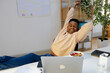 © Jimena Roquero/Stocksy - Smiling man in office stretching on his desk in front of laptop