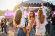 © artiemedvedev - Group of girls enjoying summer music festival on sandy beach, holding colorful drinks, smiling and dancing with stage in background. Sunlit crowd gathers around, celebrating sunny seaside event.