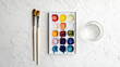 © bit - Artist's workspace with a watercolor paint palette, brushes, and a blank canvas arranged on a white textured surface, ready for creative expression