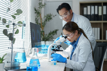 Two scientists in a laboratory setting are focused on conducting research using a microscope. The workspace is filled with laboratory equipment, including beakers and test tubes with blue liquid