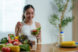 © crizzystudio - Happy young asian woman eating a healthy salad for breakfast or lunch, sitting at the table in a bright modern kitchen, enjoying fresh organic vegetables and healthy lifestyle