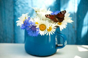 Wall Mural - a small bouquet of daisies and cornflowers with a butterfly in an old against a rustic blue wooden background with sunlight.beautiful, natural composition full of summer warmth and countryside charm.