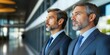 © Imagine Crane - Two men in tailored suits stand side by side in a sleek office, thoughtfully gazing ahead as sunlight streams through large windows, highlighting their focused expressions