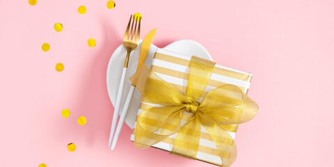 Wall Mural - Top view of heart shaped plate with gift with ribbon and golden cutlery in center of composition on pink table background. Banner.