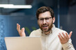 © Tetiana - Close-up portrait of a young disappointed and sad man sitting in the office at a laptop, looking at the camera and spreading his hands