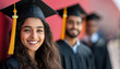 © Shivani - a young Indian woman wearing a black graduation cap and gown. Two men in the background also wear the same hats and gowns with yellow tassels on them.