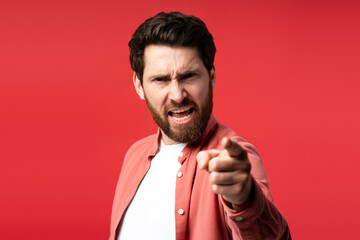 Wall Mural - Displeased bearded young man looking at camera and pointing by finger, isolated on red background