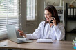 © Wasana - A woman in a white lab coat is talking on a cell phone while looking at a laptop